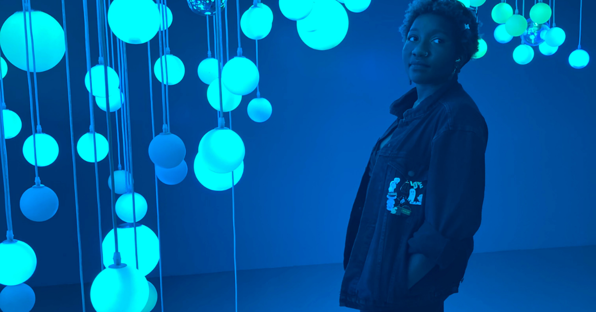 fati-banner Fati standing in a green blue room with bright teal lights hanging from the ceiling